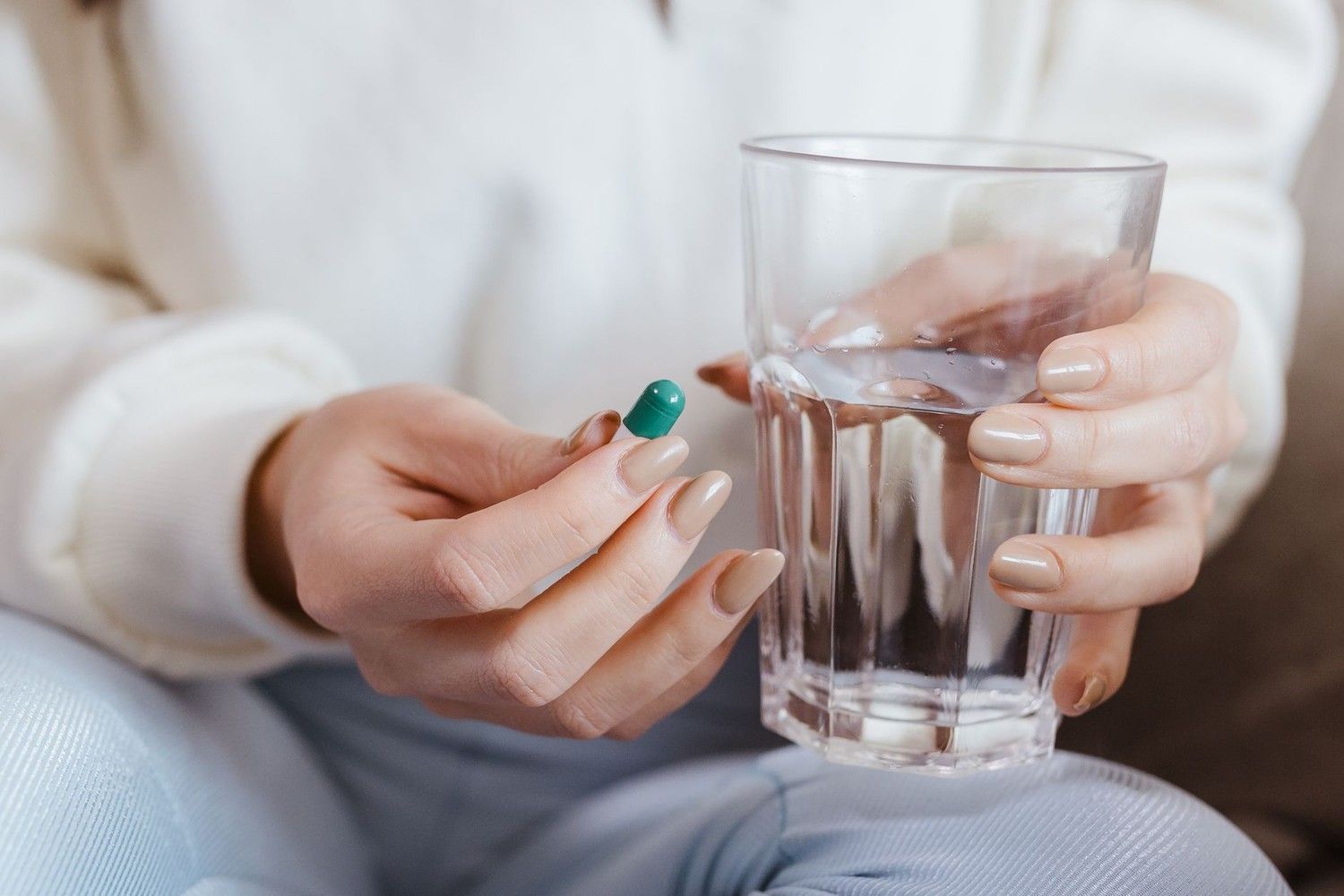 穿白色毛衣的人一手拿著綠色膠囊、一手握著一杯水