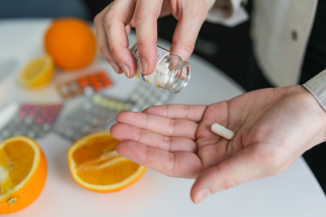 一隻手從小玻璃瓶倒出白色膠囊到掌心，桌上放著切半的柳橙與藥錠